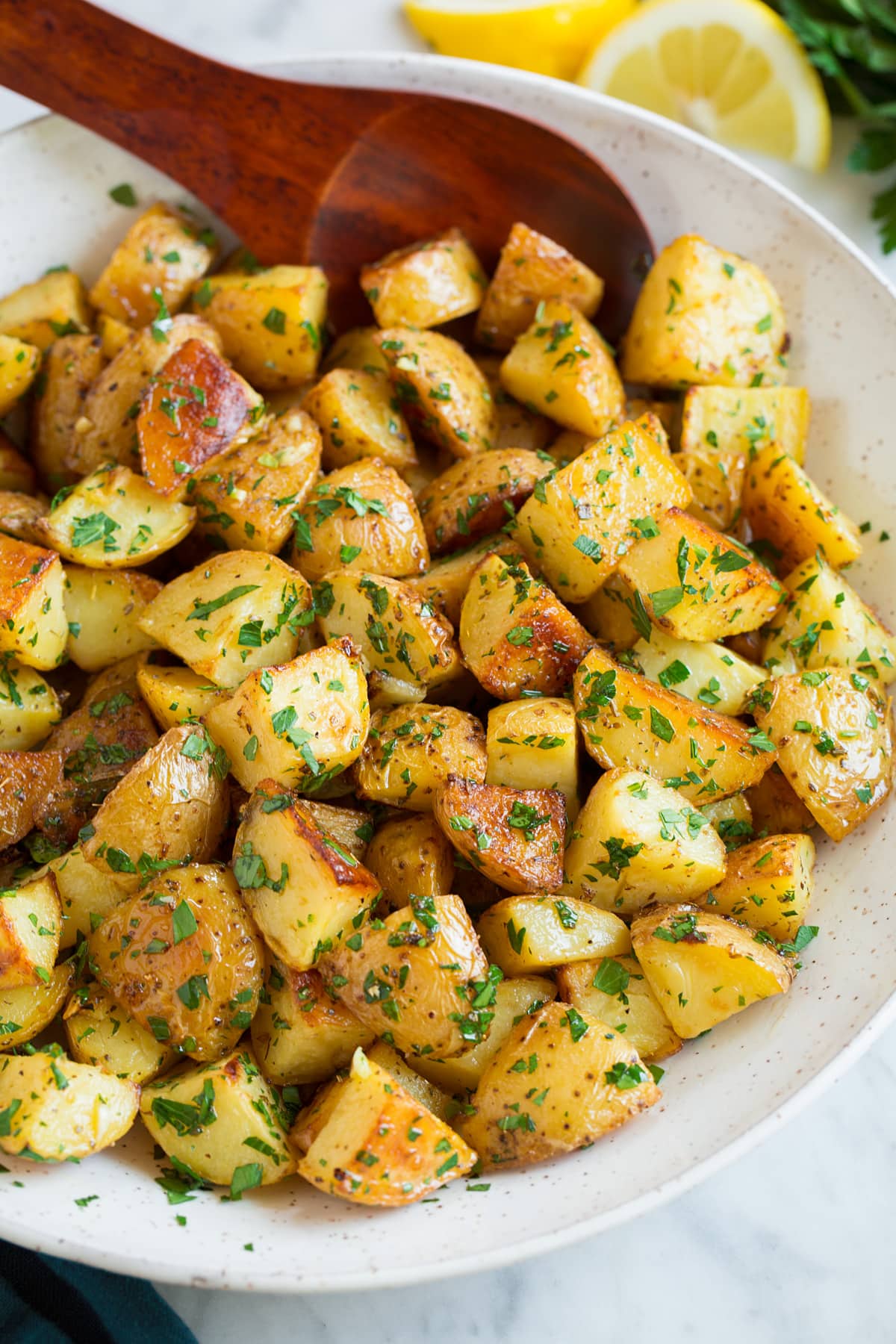 Patate Al Forno Al Limone Bowl of roasted potatoes with lemon and parsley, wooden spoon is resting in potatoes.