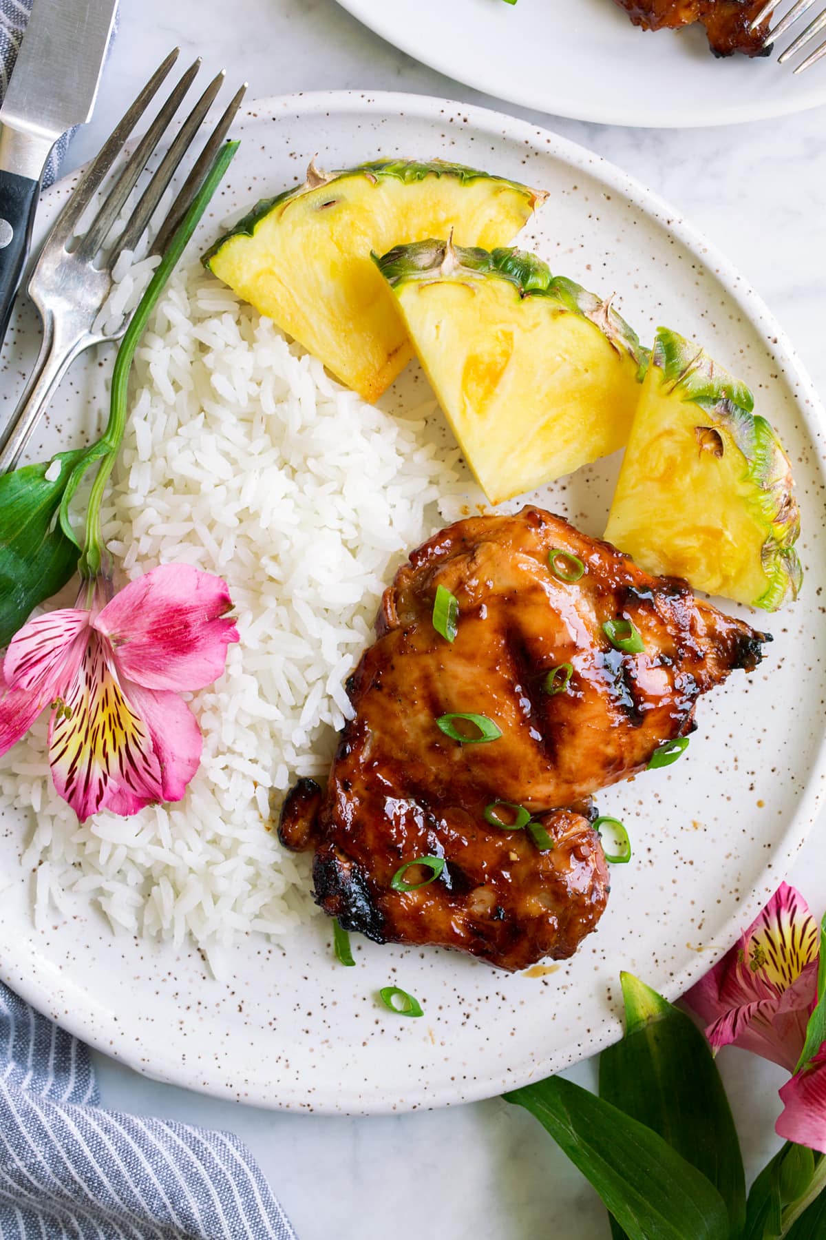Pollo Huli Huli Plate with 1 huli huli chicken piece, pineapple, rice and a fresh flower. Shown overhead.