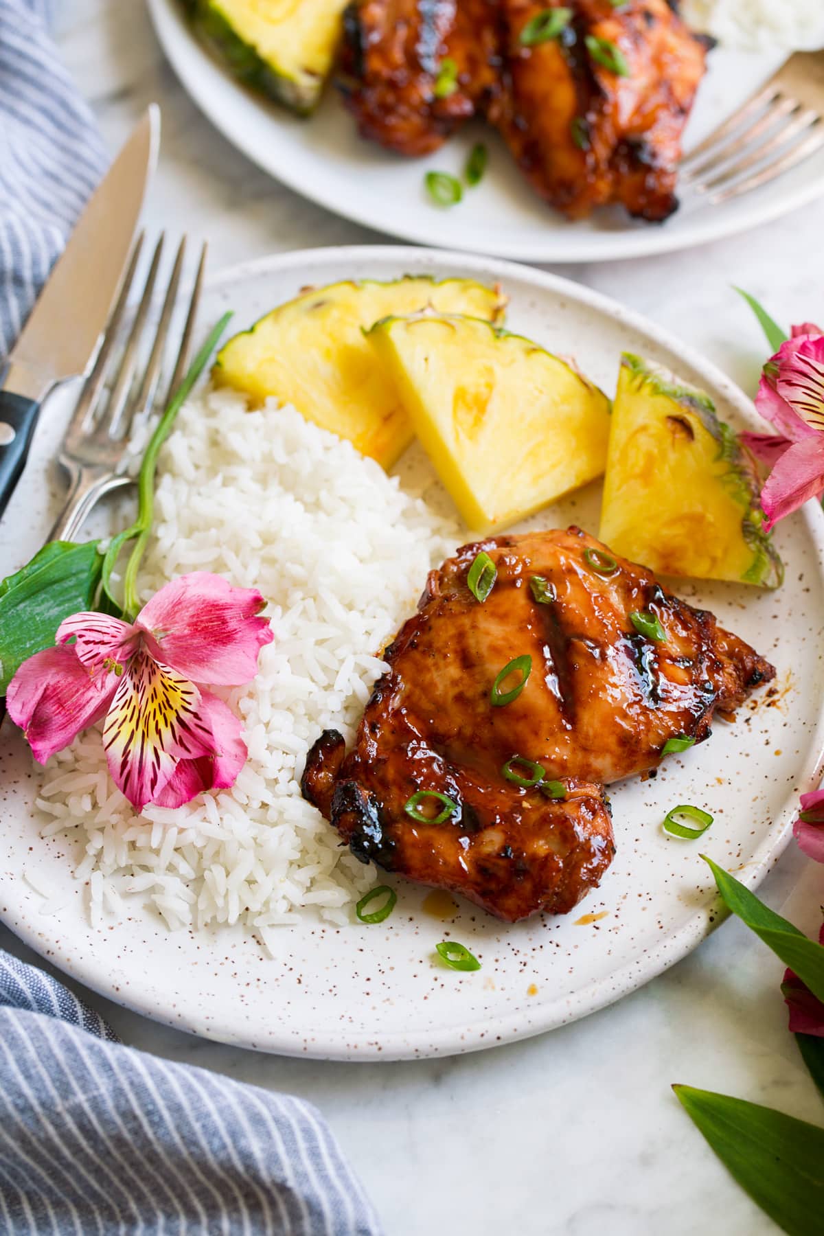 Pollo Huli Huli Plate with 1 huli huli chicken piece, pineapple, rice and a fresh flower. Shown from the side.