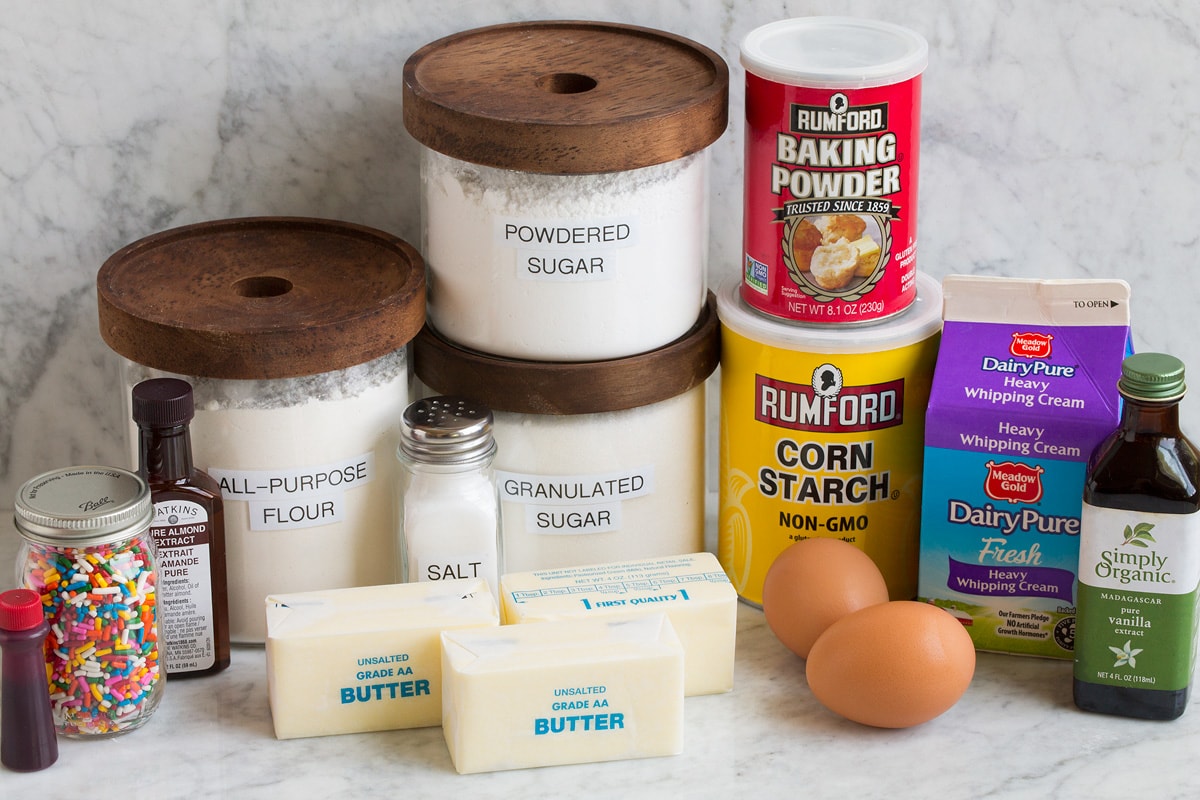 Zucchero Cookie Bar Image of ingredients that are used to make sugar cookie bars. Includes flour, sugar, powdered sugar, butter, eggs, baking powder, cornstarch, cream, vanilla, almond extract, sprinkles and food coloring.