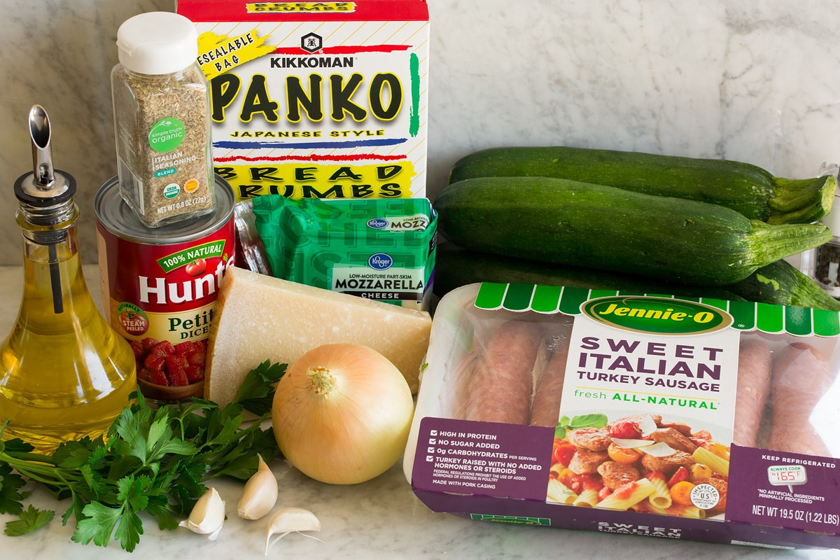 Zucchine a barchetta Image of ingredients used to make zucchini boats. Shows sausage, zucchini, onion, parmesan, parsley, garlic, mozzarella, tomatoes, Italian seasoning, panko bread crunbs and olive oil.